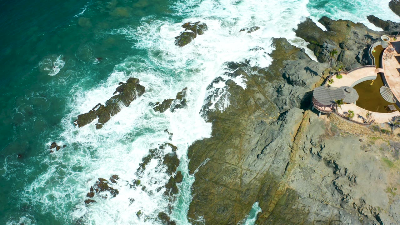 At 0:03, waves wrap around jagged Cerritos Beach rocks while a curved terrace and small pool area remain visible on the right, with no visible people.