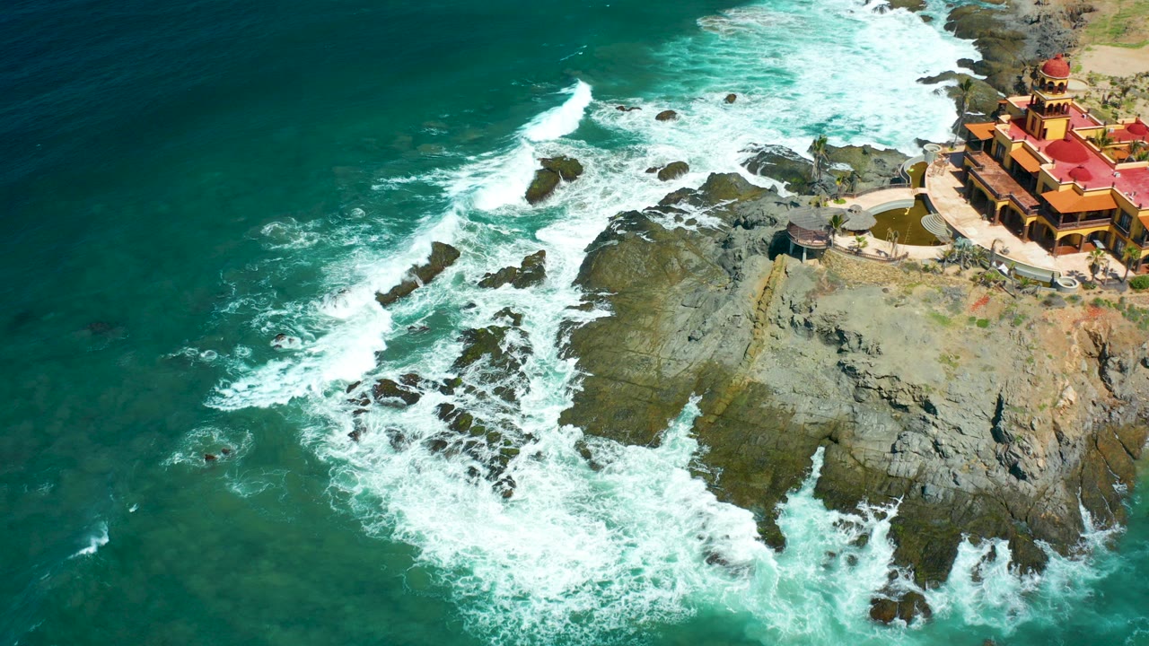 Aerial frame at Cerritos Beach, Baja California Sur shows turquoise water washing over dark rocks beside a red-roofed cliffside building, with no visible people.