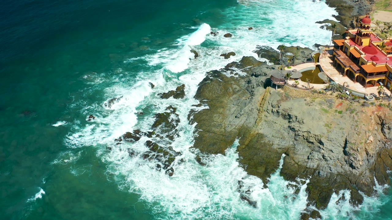 At 0:02, the aerial view over Cerritos Beach shows a bright line of breaking surf near the upper rocks and the red-roofed building partly cropped on the right, with no visible people.