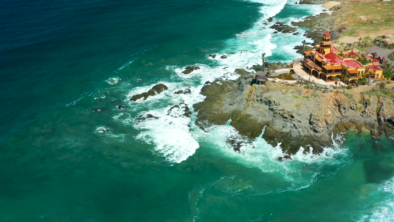 At Cerritos Beach in Baja California Sur, turquoise water and white surf surround dark rocks below a red-roofed cliffside building, with no visible people.