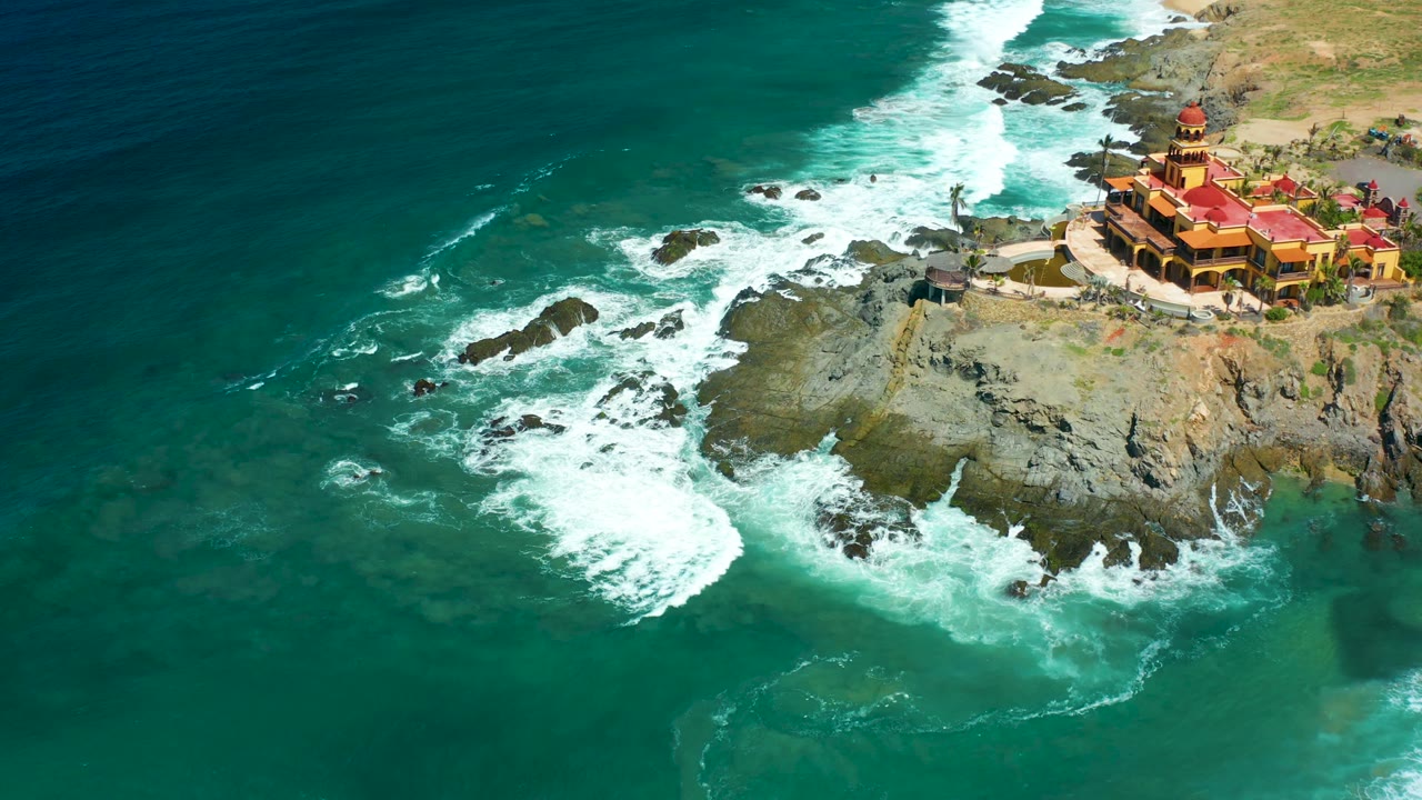 At 0:02, Cerritos Beach water shifts from deep teal to turquoise around exposed rocks beneath the red-roofed cliff building, with no visible people.
