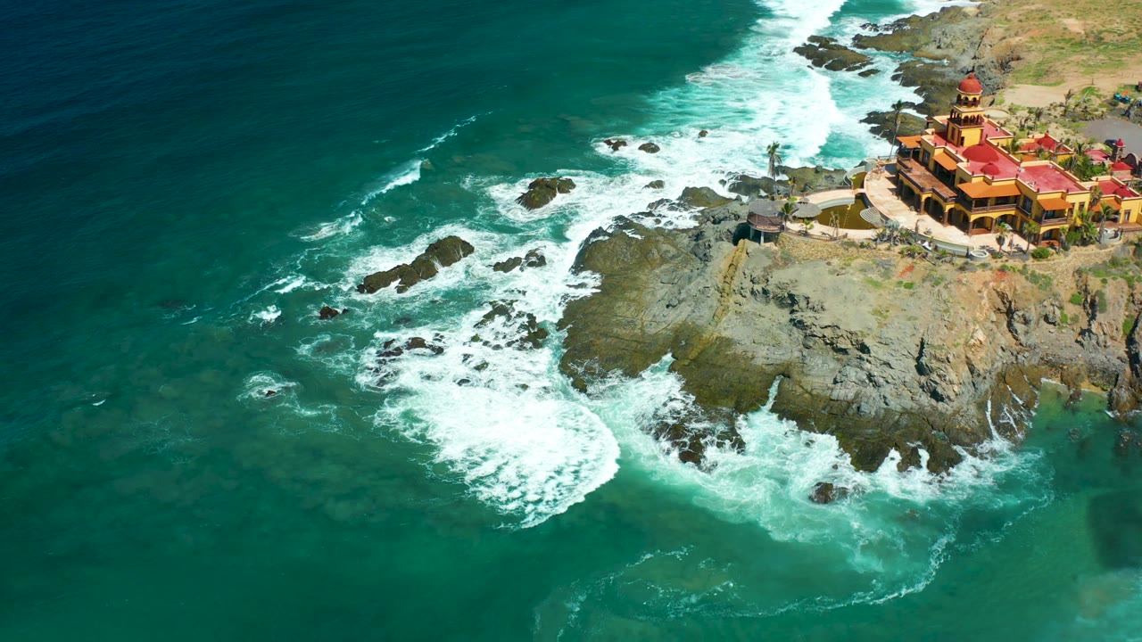 Aerial shoreline view with waves curling around rocks and a red-roofed building on the cliff.