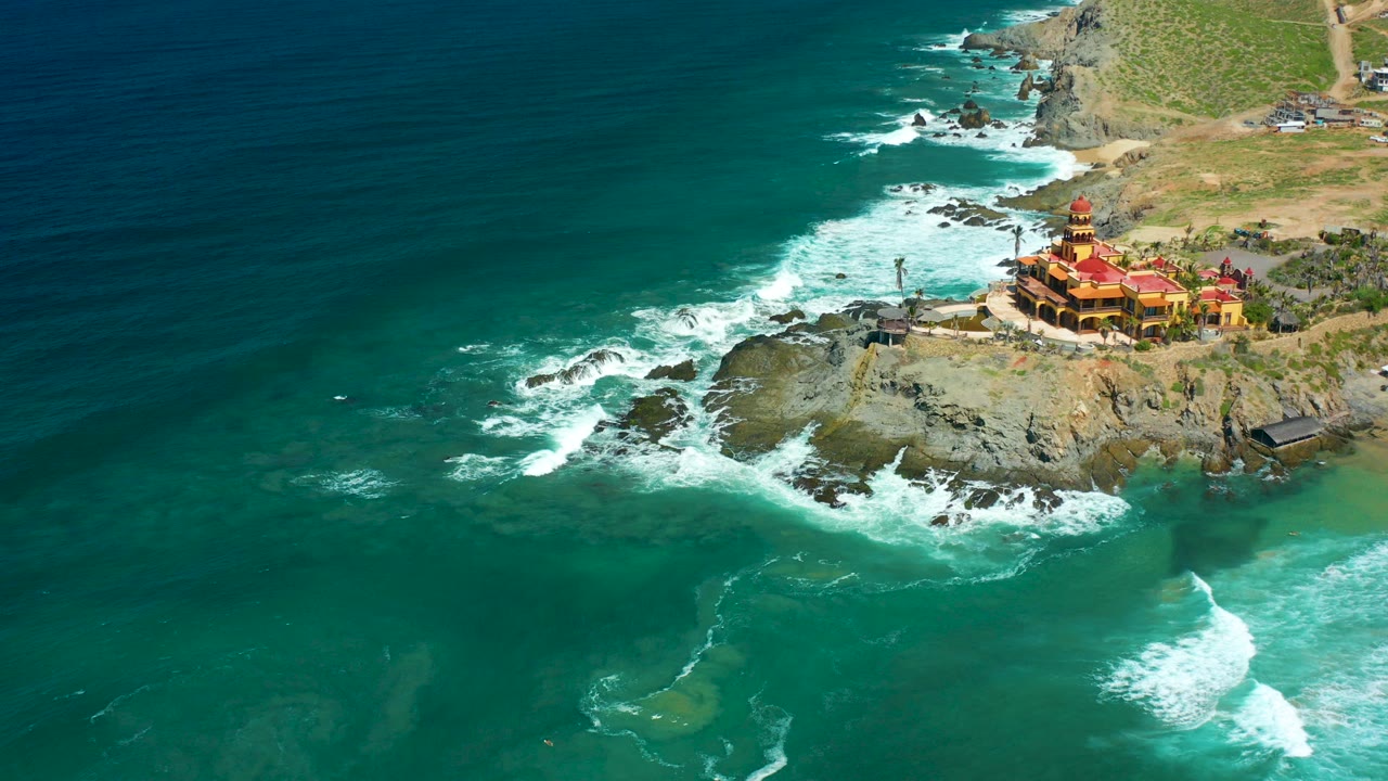 Aerial frame over Cerritos Beach, Baja California Sur, showing turquoise water, white surf around dark rocks, a red-roofed building on the right cliff, and no visible people.
