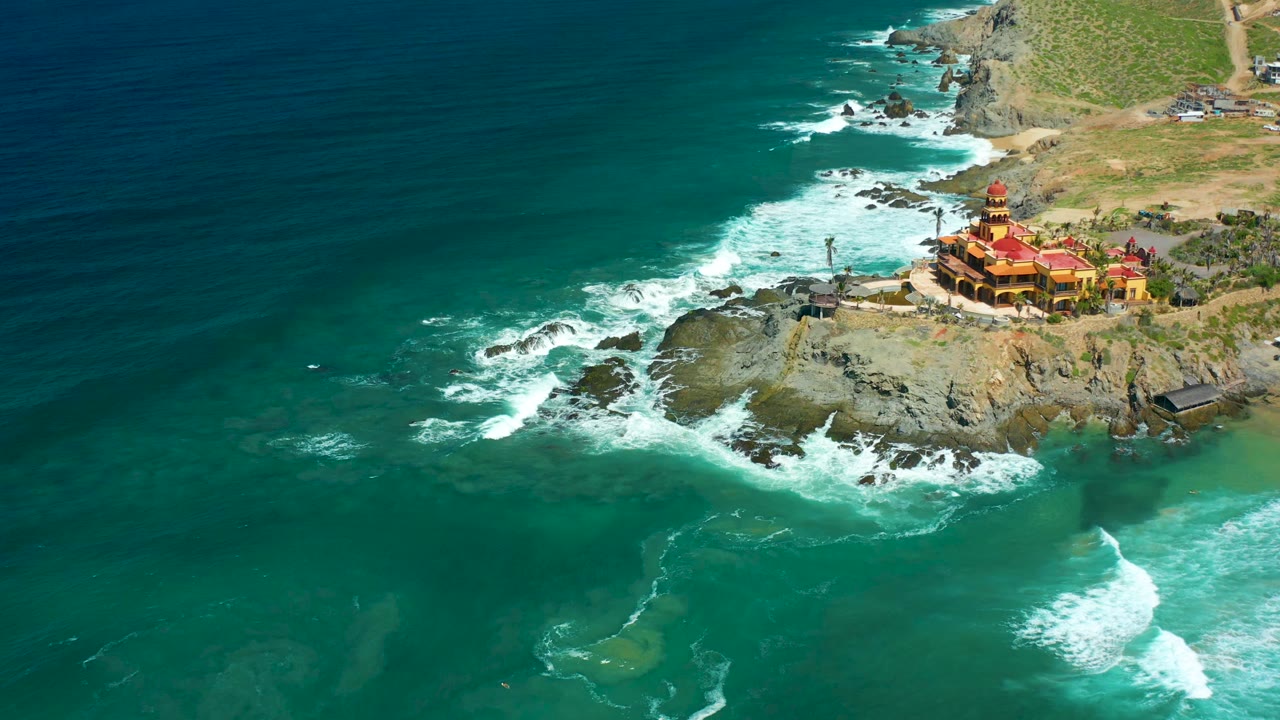 White waves break beside a rocky point below a red-roofed building.