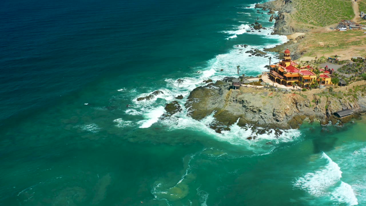 Cerritos Beach aerial frame showing a bright foam line curling through turquoise water, exposed rocks below the cliff, the red-roofed building on the upper right, and no visible people.