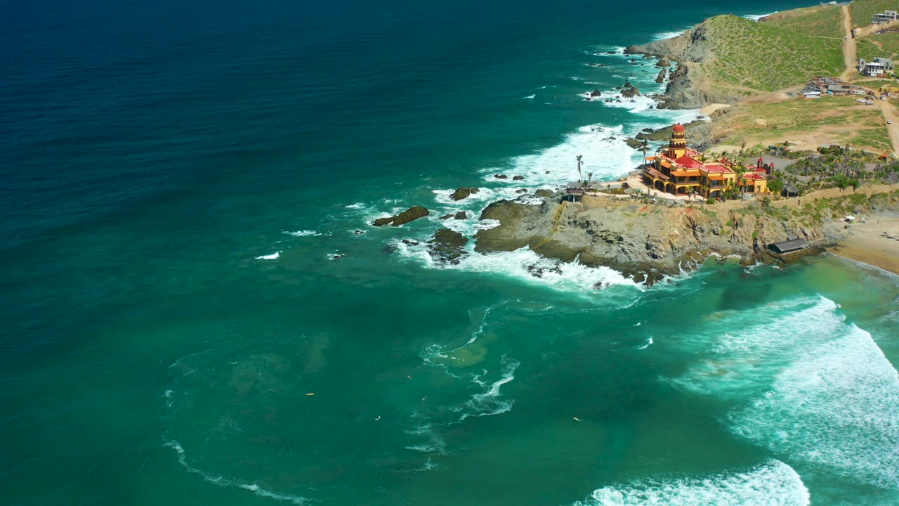 At Cerritos Beach in Baja California Sur, turquoise surf curls around rocks below a red and yellow cliffside building, with dry hillside behind it and no visible people.