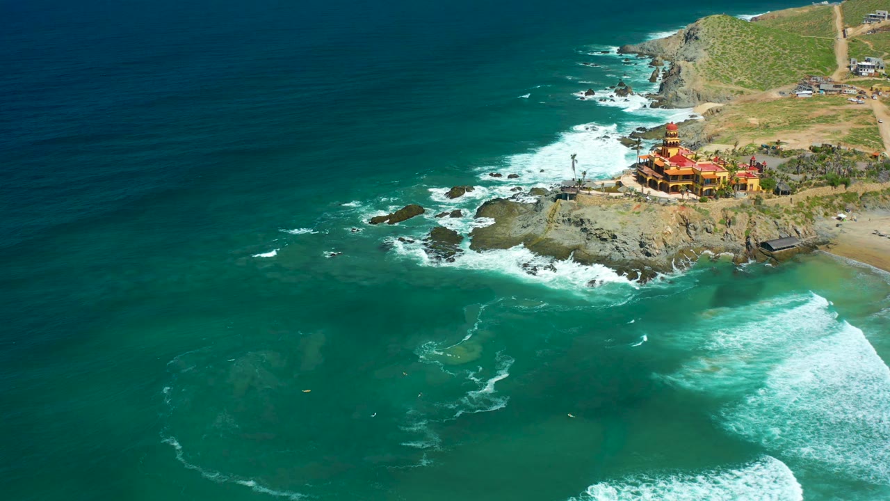 Aerial frame at Cerritos Beach shows white foam spreading across the rocks while the cliffside building stays near the upper right, with no visible people in the surf or on shore.