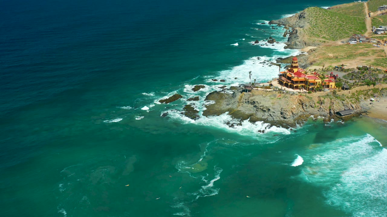 Ocean surf breaks in bands near a rocky point with a cliffside structure and hillside beyond.
