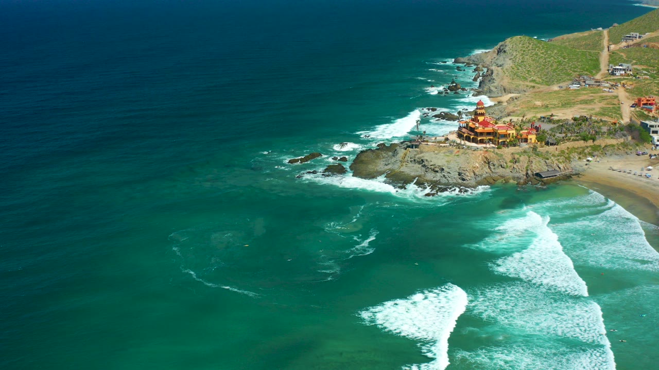 Aerial frame over Cerritos Beach, Baja California Sur, showing blue-green ocean, white surf, a rocky point, a red-and-yellow coastal structure, beach edge, hillside, and no visible people.