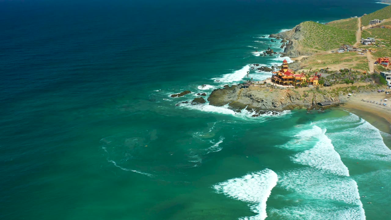 Aerial view at Cerritos Beach with more open ocean visible, white wave bands moving toward the rocky point, the cliffside coastal structure, and no visible people.