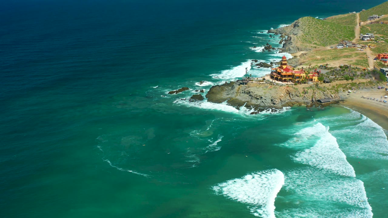 Aerial Cerritos Beach frame with the coastal structure near the upper right, turquoise water across the foreground, white foam curling over the beach, and no visible people.