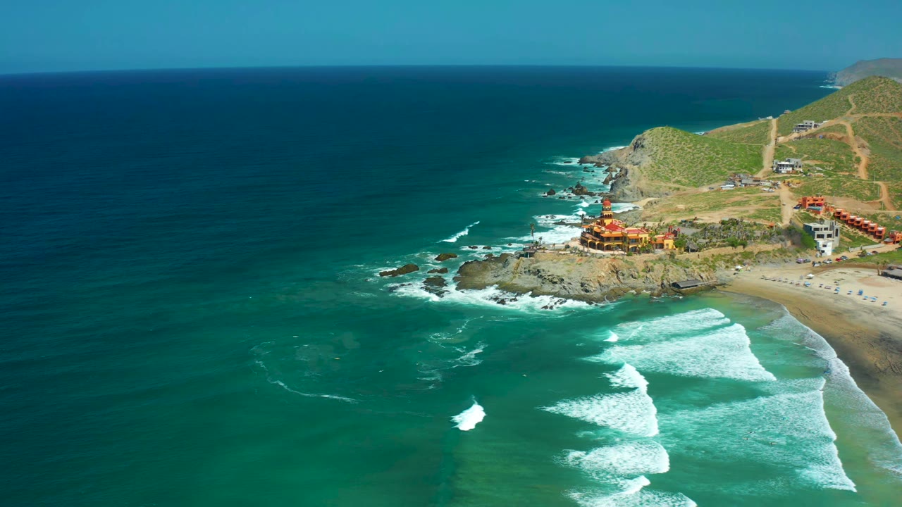 Aerial frame at Cerritos Beach, Baja California Sur, Mexico, showing surf bands moving closer to the sand, deep blue water offshore, the rocky point on the right, coastal buildings, and no visible people.