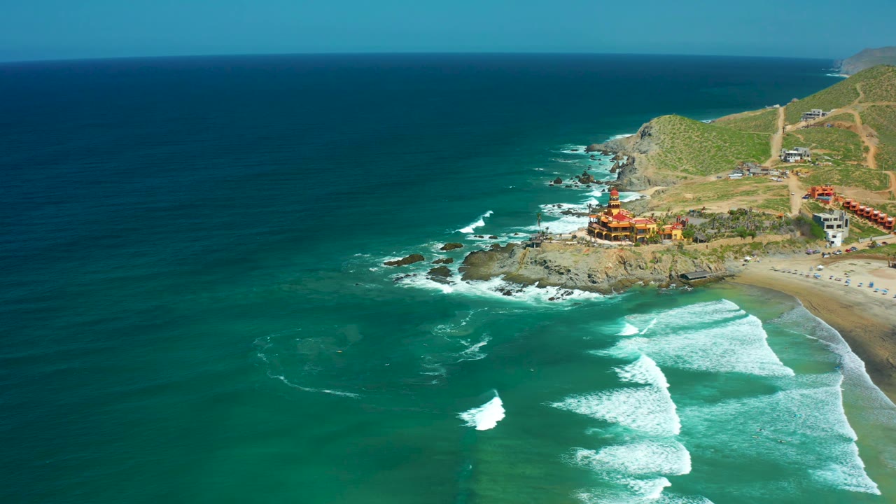 Cerritos Beach aerial frame with a larger wedge of open water on the left, white waves breaking toward the lower right beach, a rocky headland with buildings, hillside roads, and no visible people.