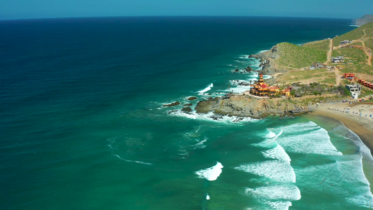 High aerial view of teal water, surf, beach, rocks, buildings, and green hills.