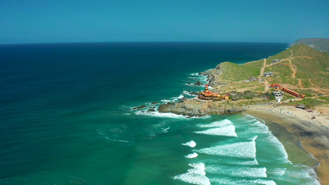Surf lines break toward the sand below a rocky coastal bluff with buildings.