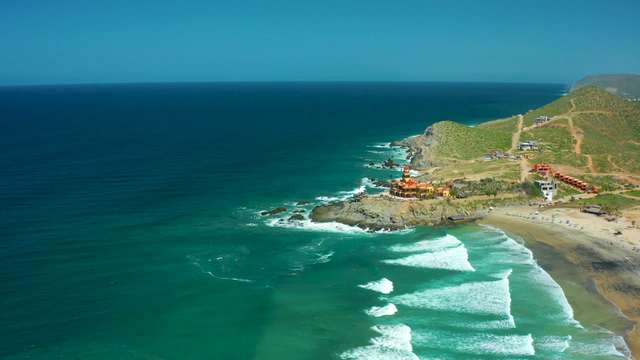 The aerial frame keeps deep blue ocean on the left, foamy waves below, Cerritos Beach sand at right, cliffside buildings above the rocks, and no visible people.