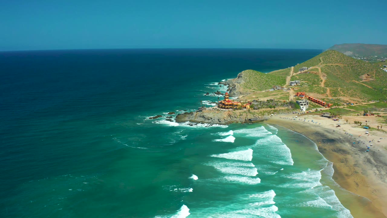 At 0:01, Cerritos Beach remains framed from above as white surf bands sit slightly farther down the shoreline, with the rocky point and hillside buildings still visible and no visible people.