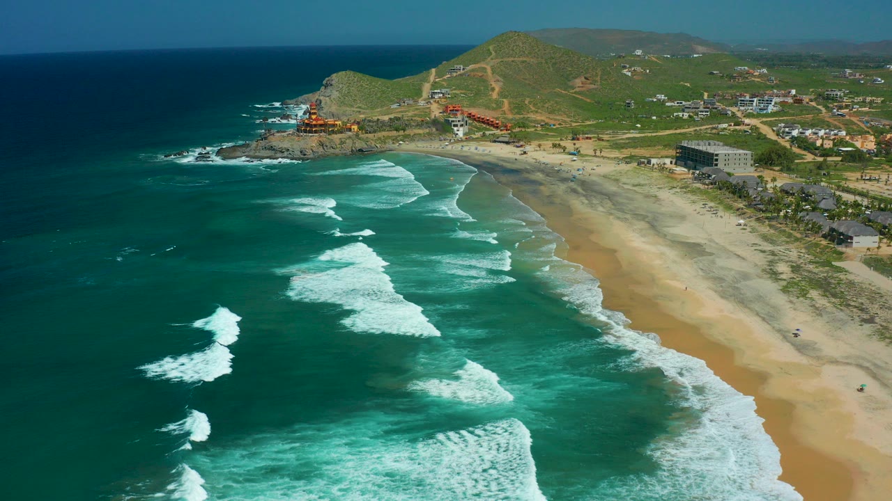 Aerial frame of Cerritos Beach in Baja California Sur with turquoise water, white surf lines, sandy shore, green hills, distant buildings, and no visible people.