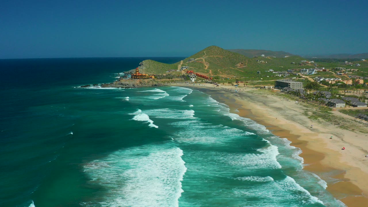 Aerial frame of Cerritos Beach, Baja California Sur, with turquoise water and white surf reaching a sandy shore, green hills and scattered coastal buildings in the background, and no visible people.
