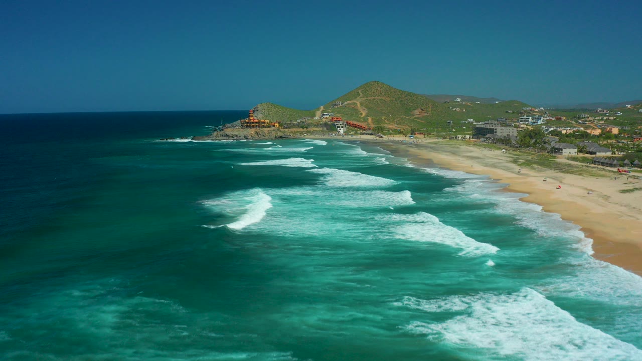 Aerial frame over Cerritos Beach, Baja California Sur, showing dark blue water, turquoise shallows, white surf bands, sandy shoreline, green hills, distant buildings, and no visible people.
