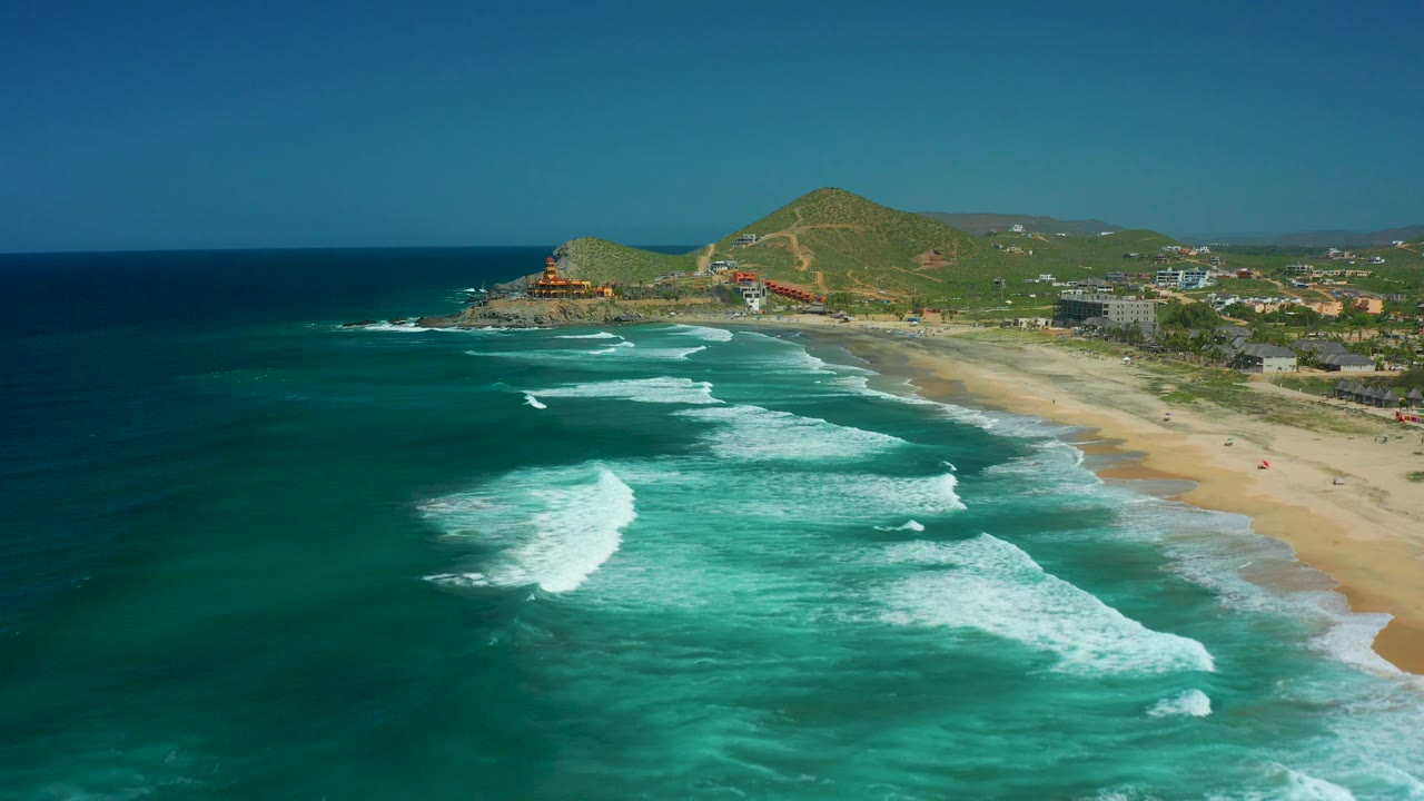 Wide drone view of surf lines approaching a sandy beach and hillside coast.