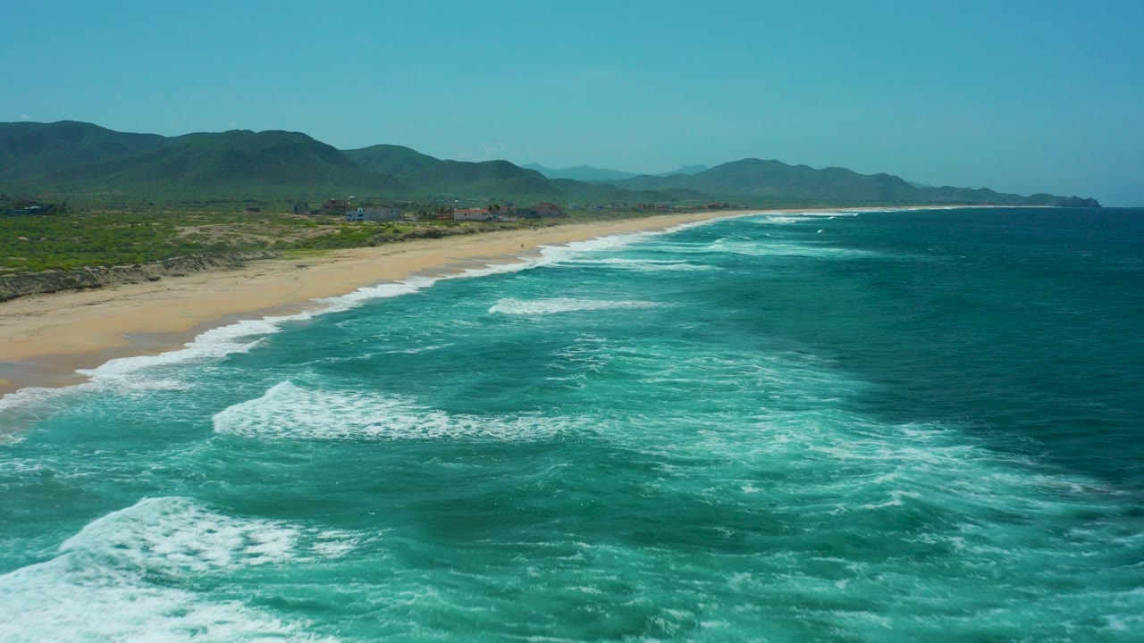 Aerial view of turquoise waves breaking beside a sandy beach and green hills.