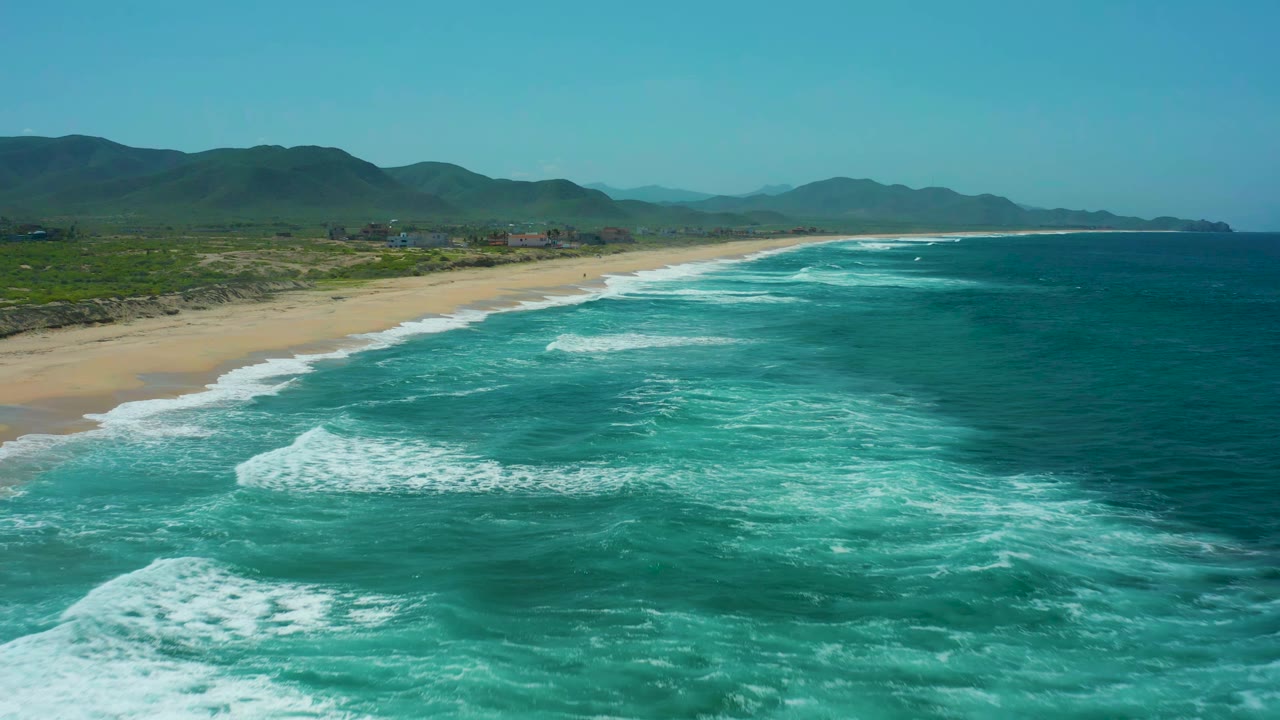 At Cerritos Beach in Baja California Sur, white surf bands widen across turquoise nearshore water while the sandy beach and green mountain backdrop remain in view, with no visible people.