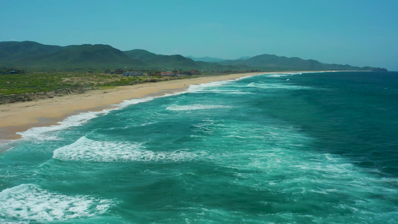 At Cerritos Beach in Baja California Sur, deeper blue ocean sits offshore while foamy turquoise water reaches the sand below green hills, with no visible people.