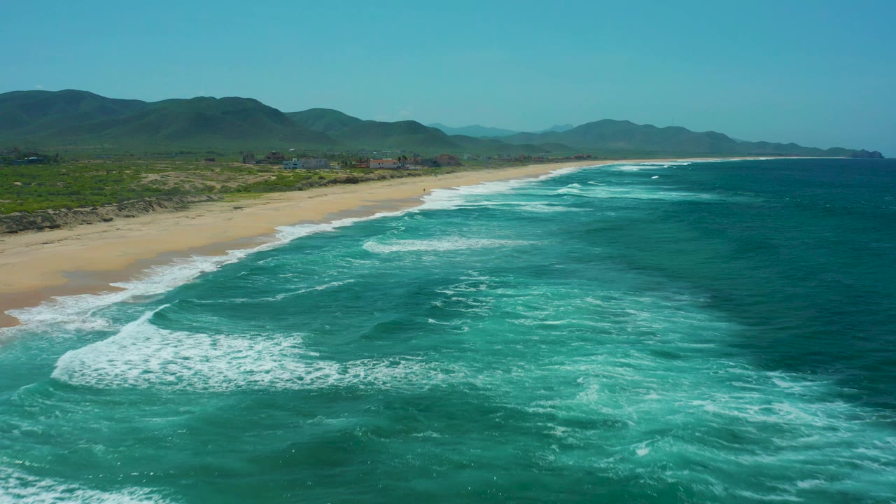 At Cerritos Beach in Baja California Sur, the breaking surf traces a diagonal line along the sandy beach, backed by green hills and a clear blue sky, with no visible people.
