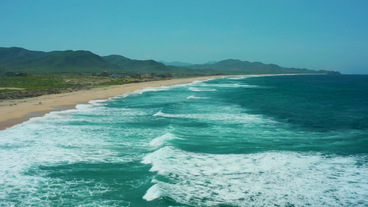Aerial view over Cerritos Beach in Baja California Sur with turquoise surf, white foam bands, sandy shoreline, green mountain backdrop, clear blue sky, and no visible people.