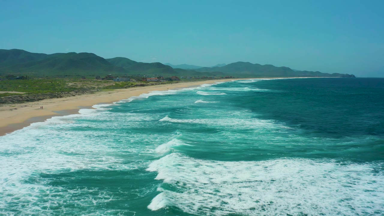 Wide aerial coastline with foamy surf rolling toward a long sandy shore.