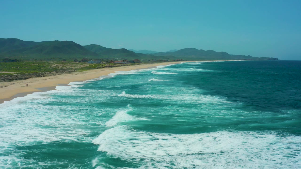 Aerial frame of Cerritos Beach with a broad turquoise water surface, surf curling toward the sand, green coastal flats, mountains behind them, and no visible people.