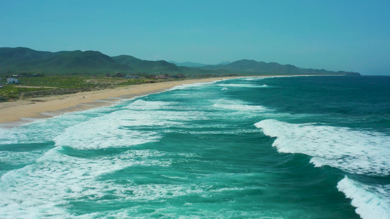 Aerial view of turquoise waves breaking beside a sandy beach and distant hills.
