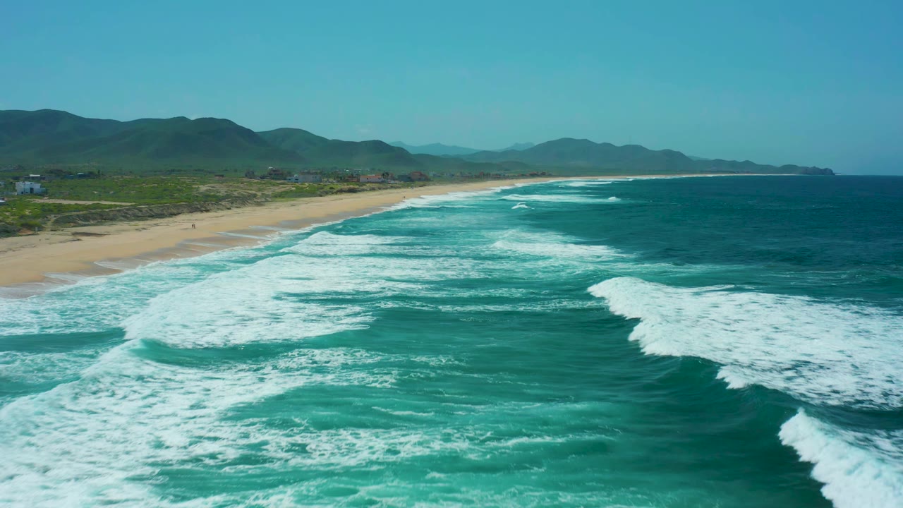 Aerial frame from Cerritos Beach showing a long sandy coast on the left, layered white breakers across the turquoise water, distant hills, blue sky, and no visible people.