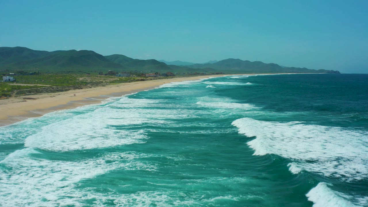 Cerritos Beach aerial frame with a broad patch of white foam across turquoise ocean, tan sand along the left edge, low green hills behind the beach, and no visible people.