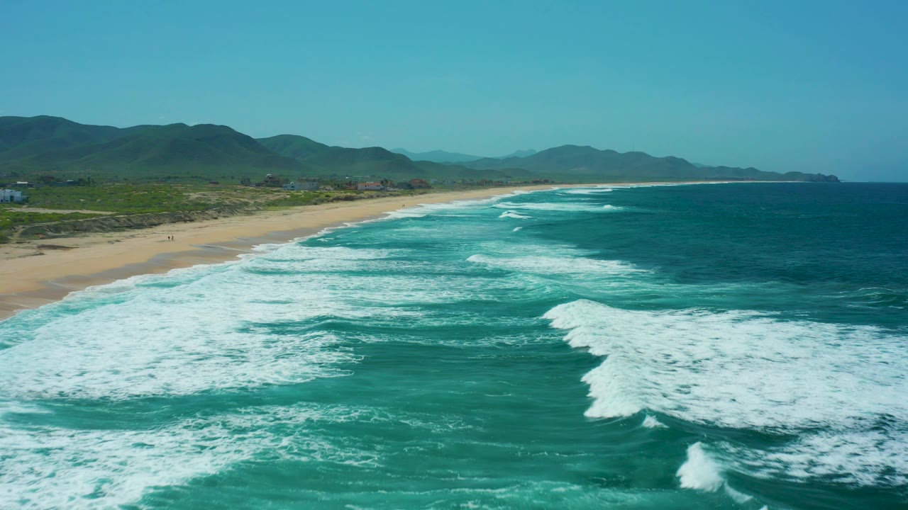 Aerial view at Cerritos Beach with whitewater filling the lower foreground, a sandy shoreline curving into the distance on the left, hills behind the coast, and no visible people.