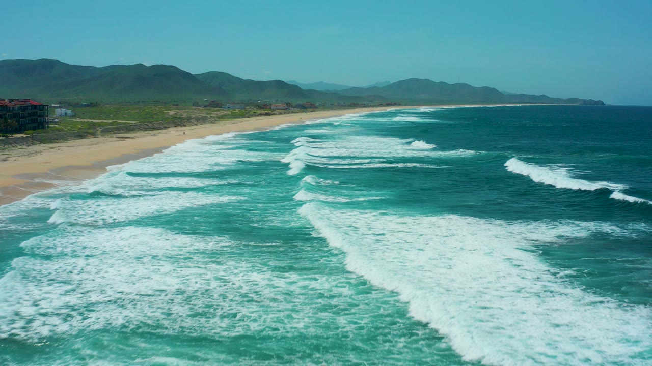 At Cerritos Beach in Baja California Sur, white surf bands fill the foreground as they approach a sandy shoreline under a clear sky, with no visible people.