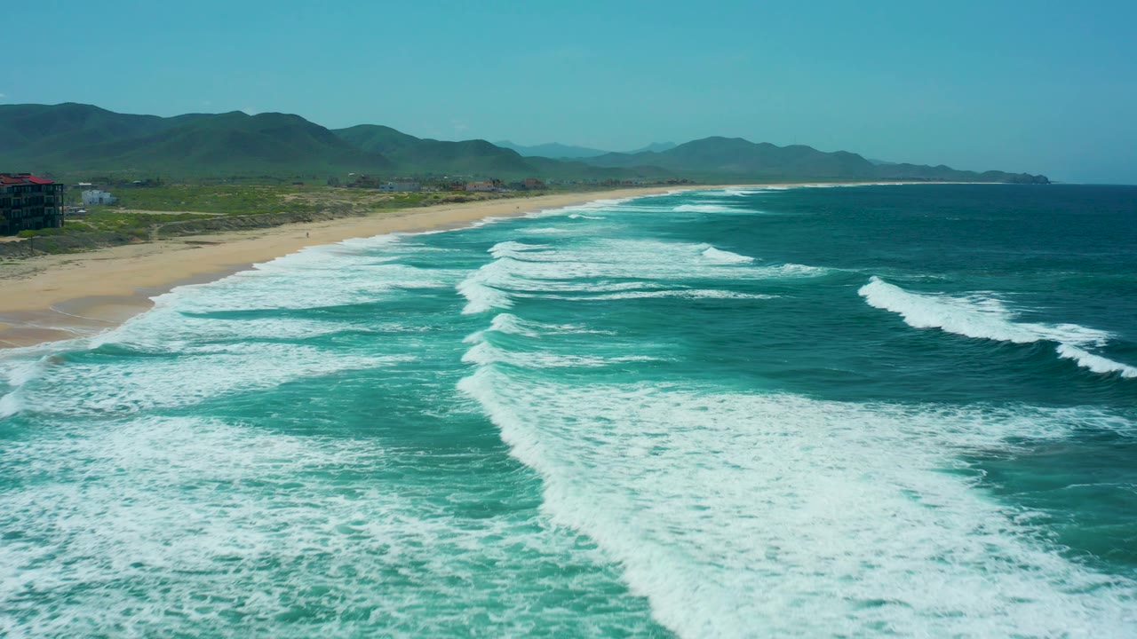 At 0:02, the foamy break sits closer to the lower frame while the long Cerritos Beach shoreline recedes toward distant mountains, with no visible people.