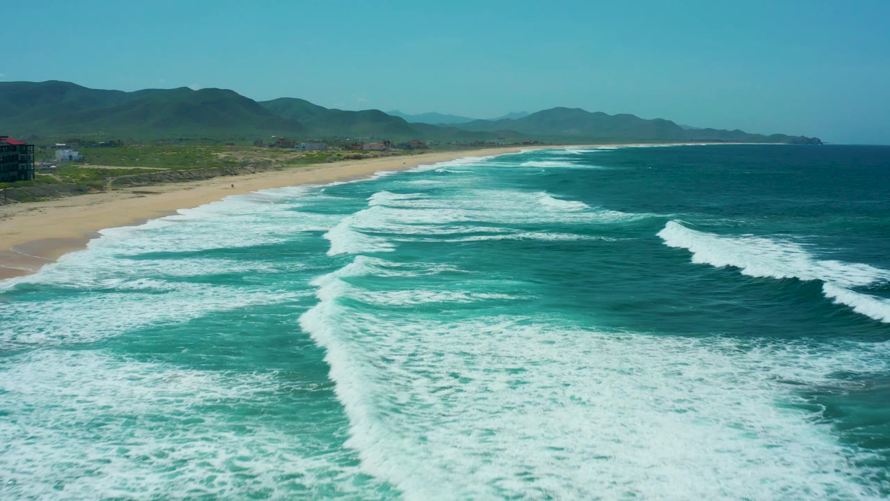 At 0:03, broad turquoise water and white foam dominate the foreground while the sandy Cerritos Beach shoreline and low mountains stay clear in the background, with no visible people.