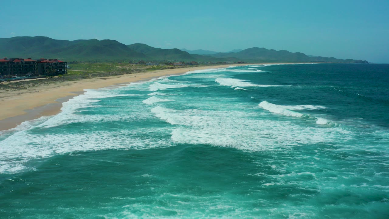 Aerial view over Cerritos Beach with turquoise surf and white foam rolling toward sand, green mountains in the distance, a beachfront building at left, and no visible people.