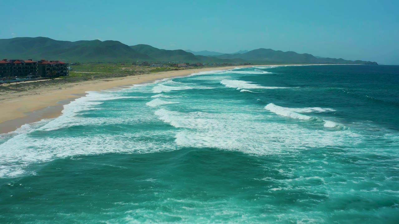 Cerritos Beach shoreline seen from above as wide white surf bands spread across turquoise water, with sand and dunes to the left and no visible people.
