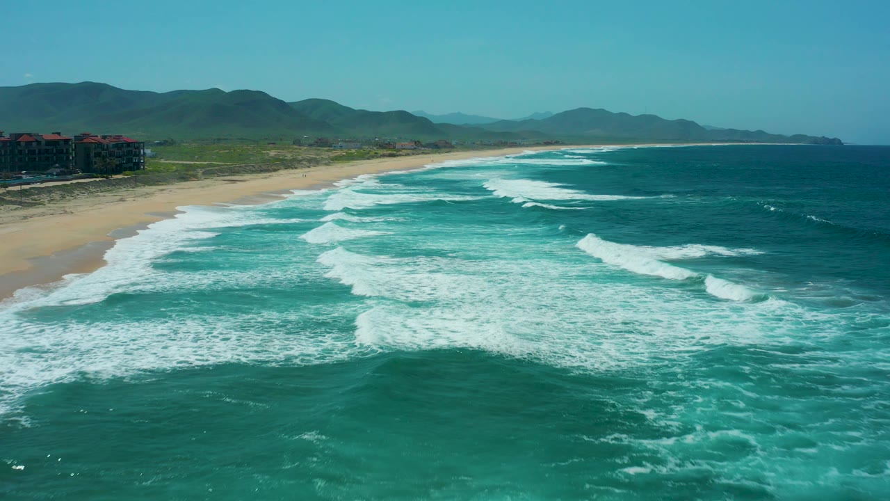 Aerial view along Cerritos Beach as the shoreline curves into the distance, with foamy waves in the foreground, darker blue ocean at right, mountains beyond, and no visible people.