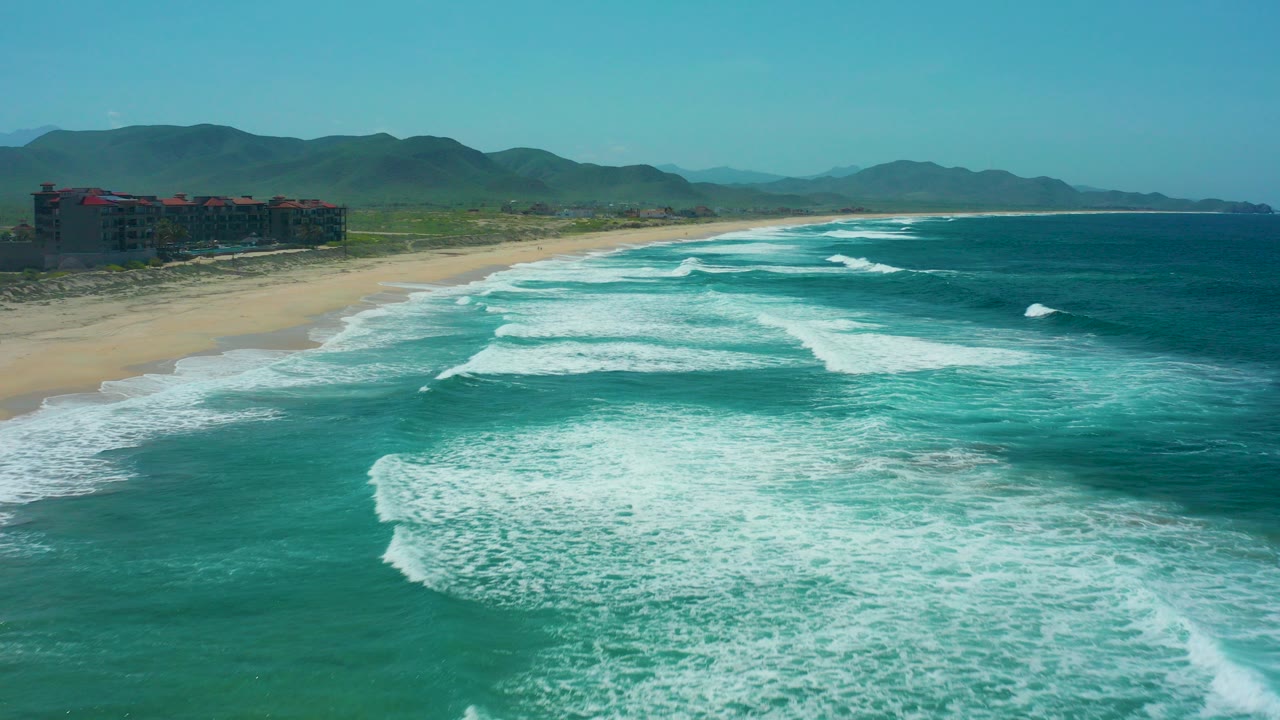 At Cerritos Beach, Baja California Sur, turquoise surf rolls toward the sandy shoreline while a beachfront building sits at the far left and no people are visible.