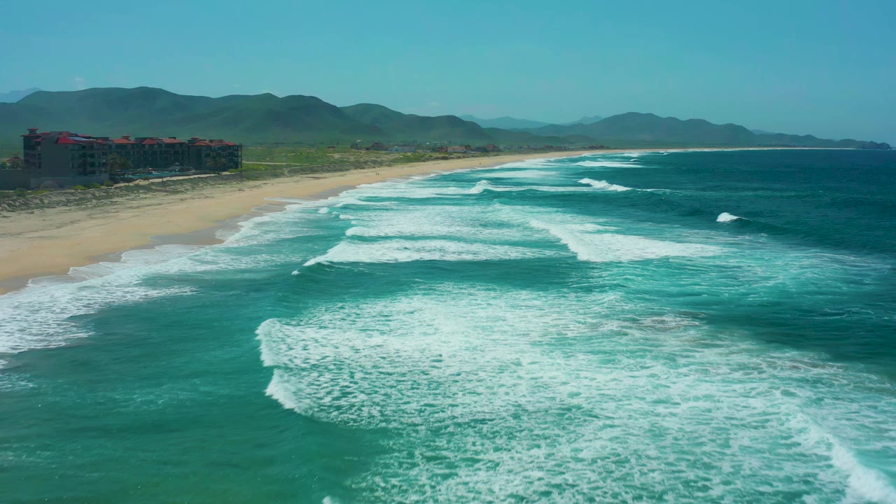 At Cerritos Beach, Baja California Sur, parallel lines of whitewater spread across the turquoise water as the sandy beach curves toward distant mountains with no visible people.