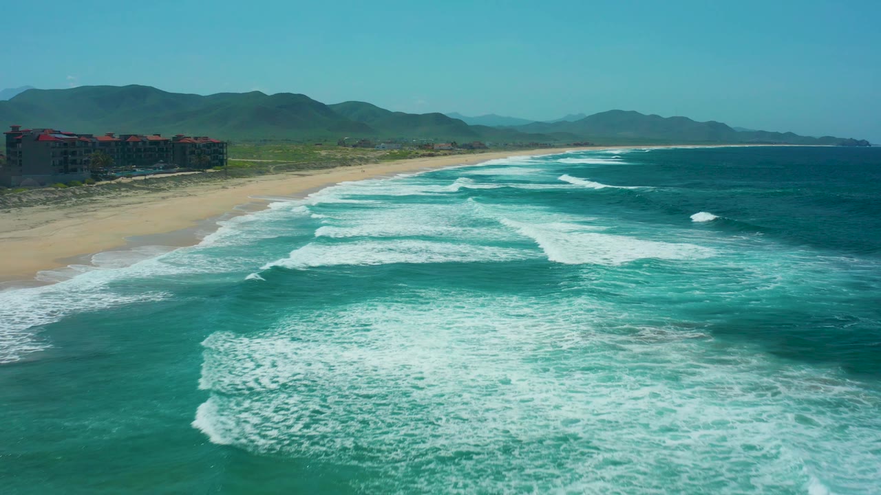 At Cerritos Beach, Baja California Sur, foamy surf fills more of the lower frame, the beach runs diagonally left to right, and the mountain ridge stays clear in the background with no visible people.