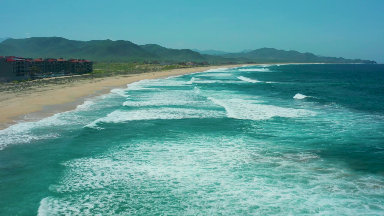 Breaking ocean waves spread white foam beside a sandy beach and mountain backdrop.