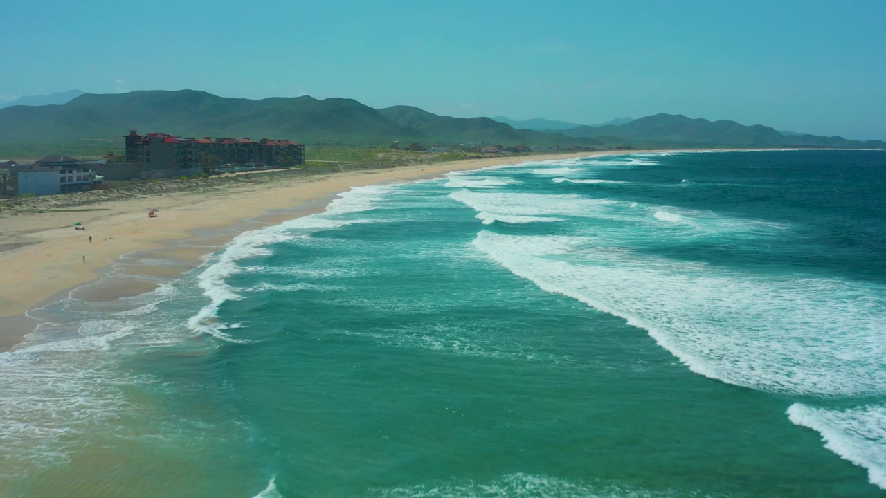 At 0:00, Cerritos Beach appears from above with turquoise surf in the foreground, a sandy shoreline on the left, coastal buildings near the beach, distant mountains, and no visible people.