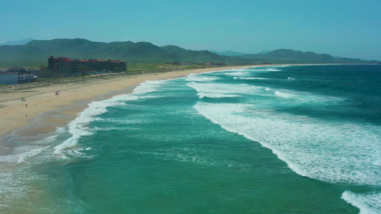 At 0:02, Cerritos Beach is framed with more open water to the right, foamy surf crossing the center, sand and coastal buildings to the left, mountains on the horizon, and no visible people.