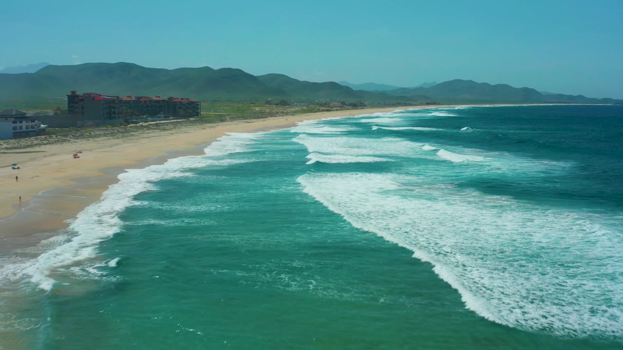 Turquoise surf curls toward a sandy beach with coastal buildings and green mountains in the distance.