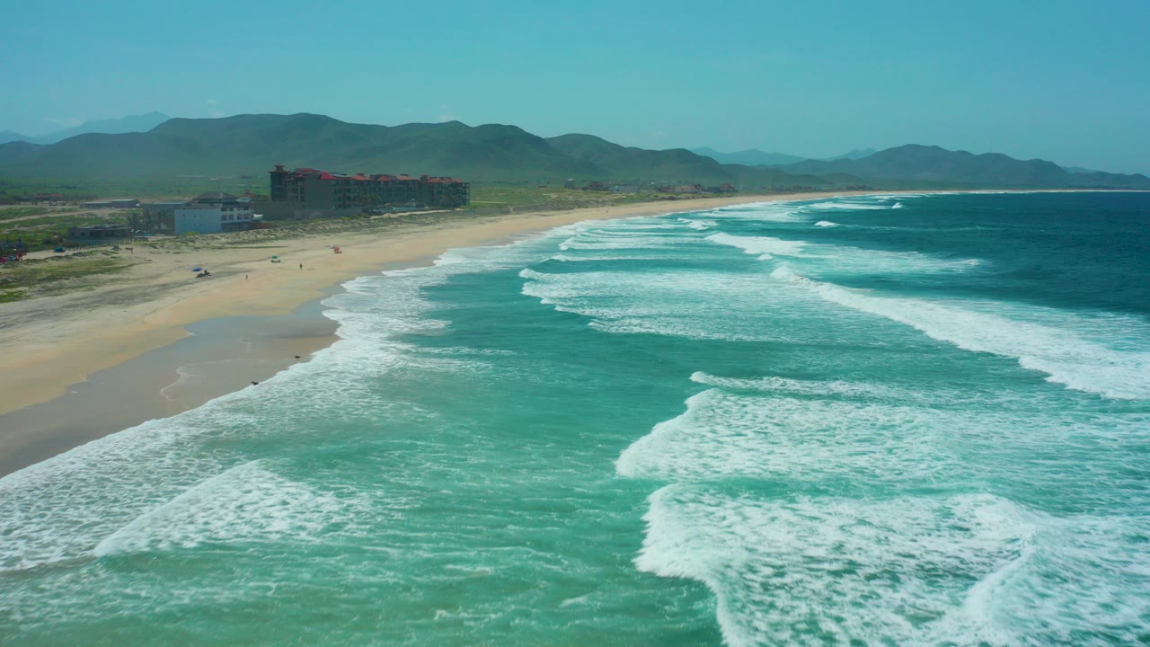 Aerial view over Cerritos Beach in Baja California Sur, with turquoise surf rolling toward a sandy shore, distant buildings at left, mountains on the horizon, and tiny people visible on the beach.